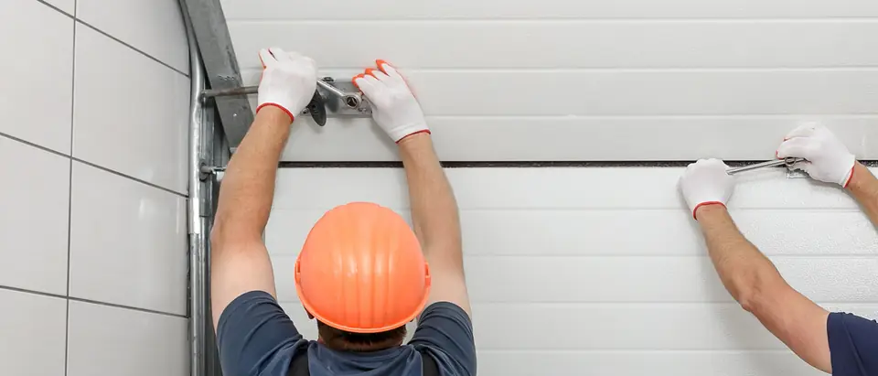 Two workers seen from behind, one in an orange hard hat, fastening hardware to the sections of a white garage door with wrenches