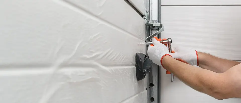Two gloved hands tightening a roller bracket with a ratchet wrench on the vertical track of a white sectional garage door