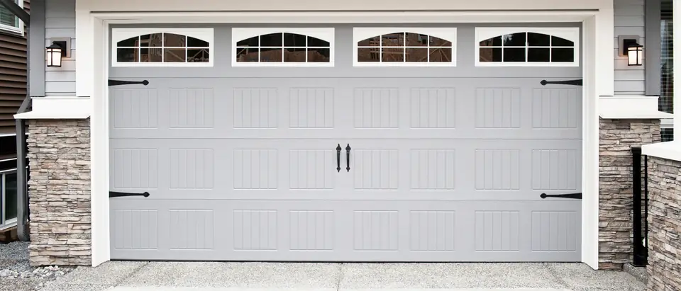 A gray two-car garage door with a row of arched windows across its top section and black strap hinges and handles, framed in white trim beside a stacked-stone column