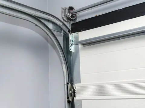 The curved steel track at the top corner of a garage door opening, with a cable drum on the shaft above and rollers in the vertical track below