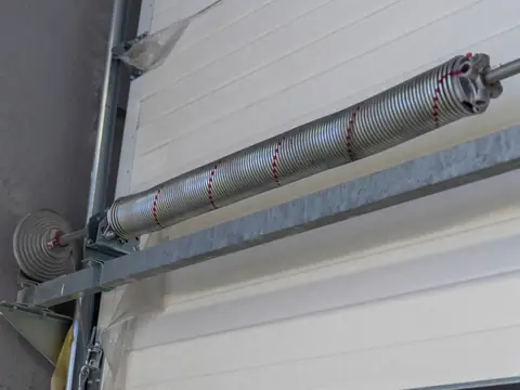 A torsion spring on a steel shaft above a white sectional garage door, with a cable drum at the end of the shaft