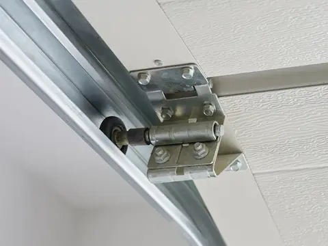A black garage door roller in its steel track, held by a hinge bolted to the edge of a door section